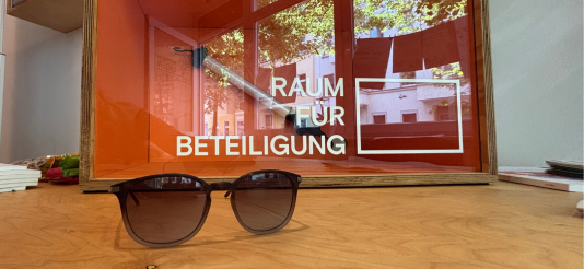 braune Sonnenbrille auf einem Holztisch hinter einem Kasten auf dem "Raum für Beteiligung" steht